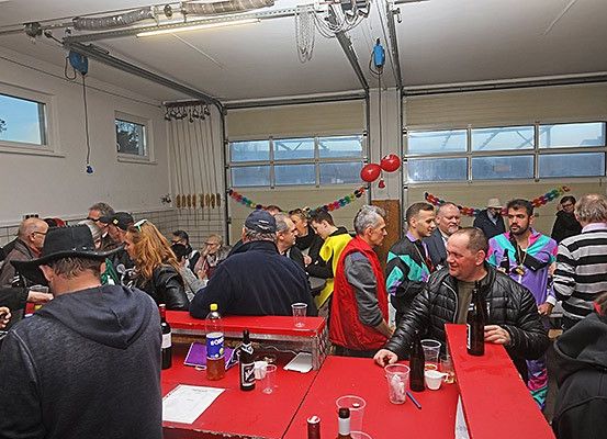Eine Gruppe von Menschen in verschiedenen Outfits, einschließlich Hüten und Jacken, versammelt sich um rote Tische in einer Garage. Sie trinken und unterhalten sich, wobei im Hintergrund Luftballons und ein Vorhang zu sehen sind.