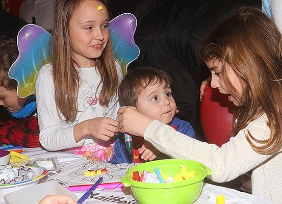 Mehrere Kinder sitzen an einem Tisch und beschäftigen sich mit einer Kunstaktivität. Ein Mädchen mit Feenflügeln hält ein kleines Objekt, während ein anderes Mädchen in eine grüne Schüssel mit kleinen Spielzeugen greift. Ein Junge hinter ihnen schaut neugierig zu.