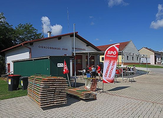 Ein Ereignis findet vor einer Feuerwache statt, mit Menschen an Tischen, einem roten Banner und Holzstapeln auf dem Boden.