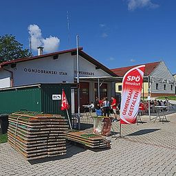 Ein Ereignis findet vor einer Feuerwache statt, mit Menschen an Tischen, einem roten Banner und Holzstapeln auf dem Boden.