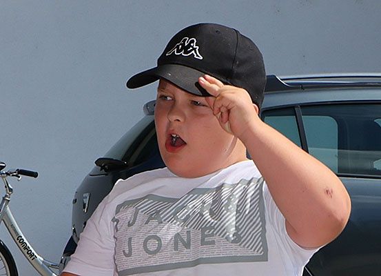 Ein junger Junge, der ein Jack Jones T-Shirt und eine schwarze Baseballkappe trägt, salutiert vor einem Fahrrad und einem Auto.