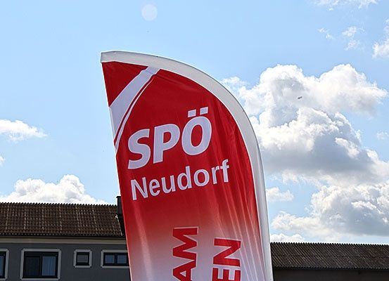 Ein roter Banner mit weißem Text 'SPO Neudorf' vor einem Gebäude unter einem teilweise bewölkten Himmel.