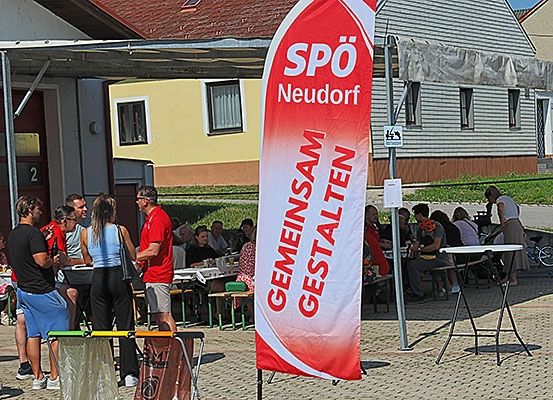 Menschen versammeln sich vor einem Gebäude, einige sitzen an Tischen. Ein roter Banner mit weißem Text lautet 'SPO Neudorf'. In der Nähe ist eine Person mit einem Fahrrad zu sehen.