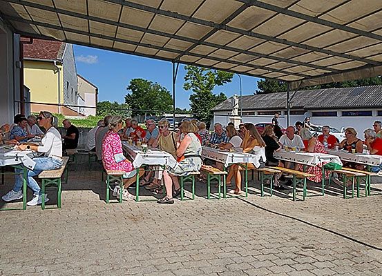 Eine Gruppe von Menschen sitzt unter einem Vordach an einem Tisch und isst. Die Tische sind mit weißen Tischdecken bedeckt. Die Umgebung ist im Freien, mit Gebäuden und Bäumen im Hintergrund.