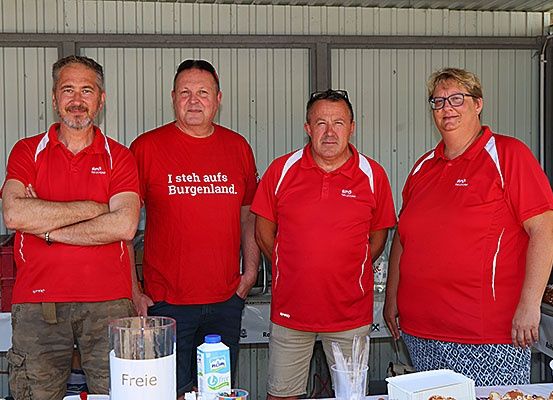 Vier Erwachsene in roten T-Shirts stehen zusammen. Drei sind Männer, eine ist eine Frau. Sie lächeln. Hinter ihnen steht ein Tisch mit Essen und Getränken.
