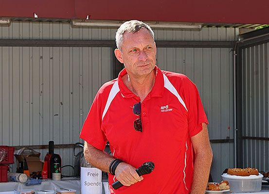 Ein erwachsener Mann in einem roten Shirt steht in einem Schuppen und hält ein Mikrofon. Hinter ihm befindet sich ein Tisch mit Essen und Getränken.