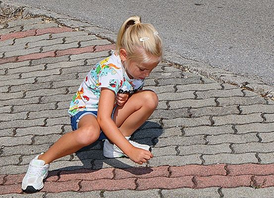 Ein kleines Mädchen hockt sich auf einen gepflasterten Gehweg und schaut auf ihre weißen Turnschuhe. Sie trägt ein farbenfrohes T-Shirt und blaue Shorts.