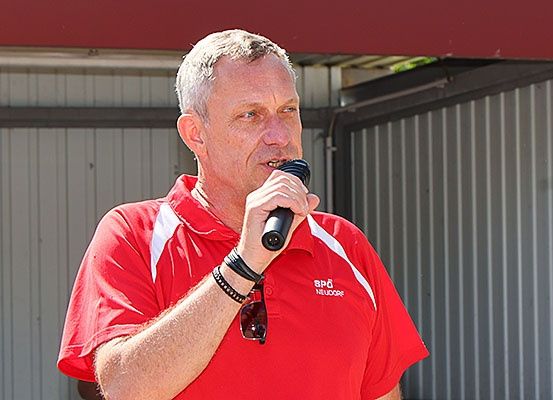 Ein Mann in einem roten Poloshirt steht draußen und hält ein Mikrofon, wahrscheinlich bei einer Rede.