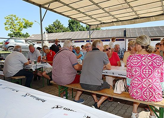 Eine Gruppe von Menschen sitzt an Bänken unter einem Zelt, einige tragen Brillen und Sonnenbrillen. Tische sind mit weißen Tüchern bedeckt, und ein Fahrrad ist in der Nähe geparkt. Im Hintergrund befinden sich Gebäude und Bäume.