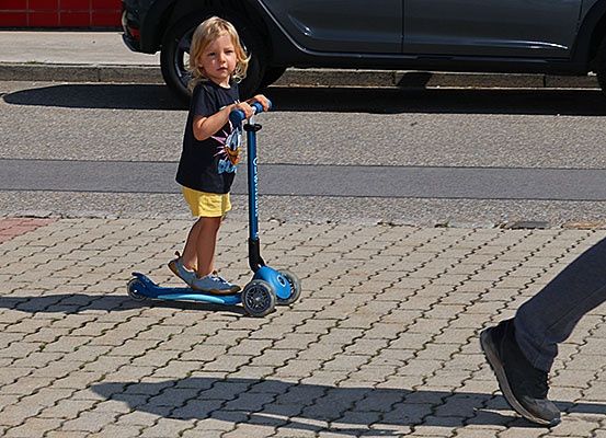 Ein kleines Mädchen in einem schwarzen T-Shirt und gelben Shorts fährt einen blauen Roller auf einer gepflasterten Straße, mit einem geparkten Auto im Hintergrund.