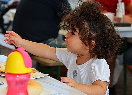 Ein junges Kind mit lockigem Haar sitzt an einem Tisch und greift nach einem Sippy-Becher. Ein Sandwich und Brot liegen vor ihm auf dem Tisch. Im Hintergrund sitzen zwei Personen an einem Tisch.