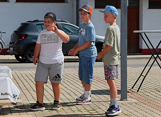 Drei Jungen stehen vor einem Gebäude, zwei mit Mützen, einer lacht. Hinter ihnen steht ein Tisch, ein Fahrrad und ein geparktes Auto.