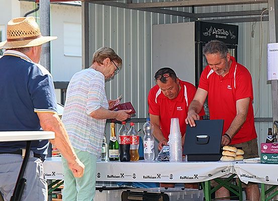 Drei Personen stehen an einem Tisch im Freien. Zwei Männer in roten Hemden bereiten Essen zu, während eine Frau in einem weißen Hemd einen Geldbeutel untersucht. Flaschen und eine Schachtel stehen auf dem Tisch.