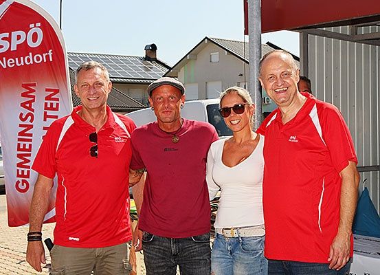 Drei Menschen in roten Shirts und eine in einem weißen Shirt lächeln für die Kamera im Freien. Hinter ihnen steht ein Banner mit 'SPO' und 'Neudorf' in Rot. Im Hintergrund sind Häuser mit Solarpanelen zu sehen.