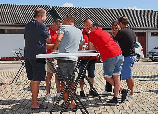 Eine Gruppe von Menschen ist um einen Tisch draußen versammelt. Einige tragen rote Hemden, andere blaue. Sie scheinen in Gespräche vertieft zu sein. Ein Gebäude mit einem gefliesten Dach und ein geparktes Auto sind im Hintergrund zu sehen.