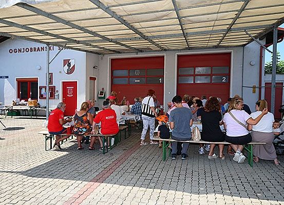 Menschen sitzen um Tische herum unter einem Schutzdach, in der Nähe eines Gebäudes mit roten Türen und einem Schild, das 'OGNJOBRANSKI' besagt. Einige Personen tragen Helme und Turnschuhe.