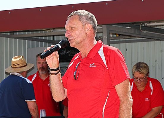 Ein Mann in einem roten Shirt spricht in ein Mikrofon bei einer Outdoor-Veranstaltung, mit zwei Personen in roten Shirts hinter ihm, eine trägt eine Brille und die andere einen Hut.
