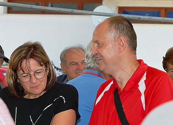 Eine Gruppe von Menschen steht und lächelt. Eine Frau mit Brille schaut nach unten. Ein Mann mit einem roten Poloshirt lächelt und schaut die Frau an.
