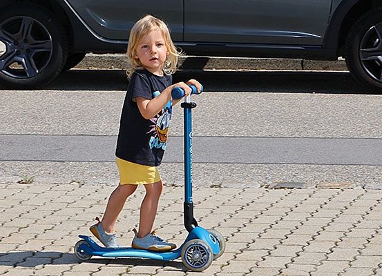 Ein kleines Kind fährt einen blauen Roller auf einem gepflasterten Bürgersteig, mit einem geparkten Auto dahinter.