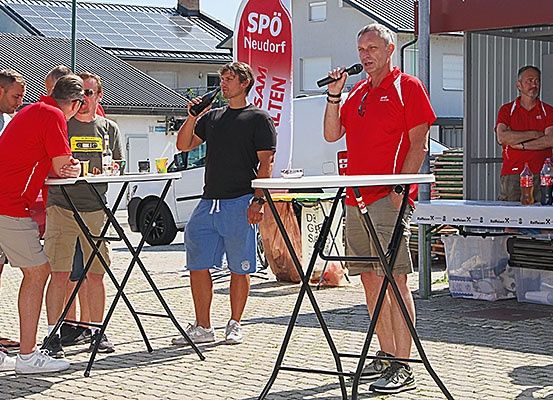 Eine Gruppe von Männern steht draußen in der Nähe von Tischen, ein Mann spricht in ein Mikrofon, ein anderer hält eine Flasche. Hinter ihnen steht ein weißer Lieferwagen und ein Gebäude mit Solarmodulen auf dem Dach.