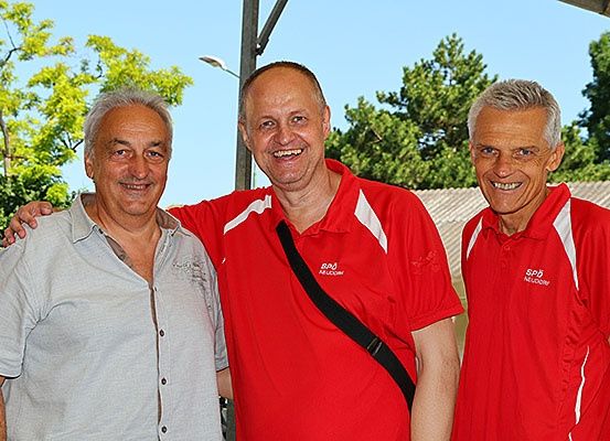 Drei Männer in roten Shirts stehen nebeneinander und lächeln. Einer hat eine Tasche. Im Hintergrund sind Bäume und eine Straßenlampe zu sehen.