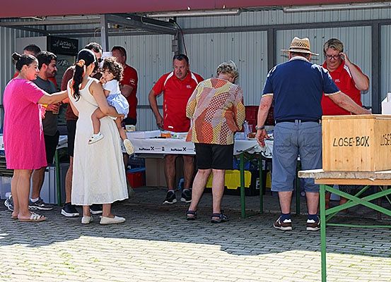 Eine Gruppe von Menschen steht um einen Tisch herum, möglicherweise bei einer Veranstaltung. Eine Frau hält ein Baby, während eine andere Frau ein Glas hält. Zwei Männer in roten Hemden stehen in der Nähe eines Tisches.