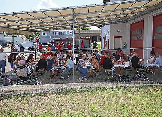 Eine Gruppe von Menschen sitzt unter einem Baldachin vor einer Feuerwache zum Essen. Viele tragen rote T-Shirts, und in der Nähe sind Fahrräder geparkt. Gebäude und geparkte Autos sind im Hintergrund zu sehen.