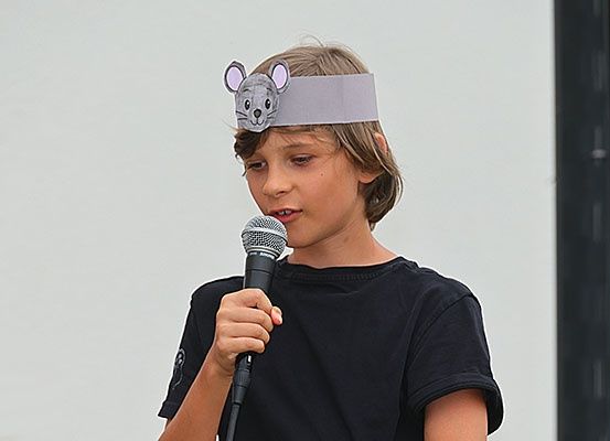Ein junger Junge mit einer Maus-Kopfbinde spricht in ein Mikrofon. Er trägt ein schwarzes T-Shirt.