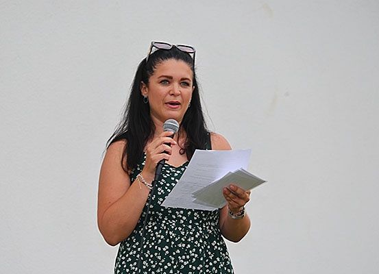 Eine Frau mit dunklem Haar spricht in ein Mikrofon und hält ein Stück Papier in der Hand. Sie trägt ein grünes Blumenkleid und hat eine Brille auf dem Kopf.