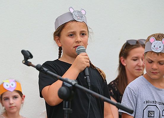 Ein Mädchen singt in ein Mikrofon, während es eine Mauskopfbedeckung trägt. Hinter ihr schauen ein weiteres Kind mit einer Mauskopfbedeckung und eine Frau mit einer Sonnenbrille auf sie.