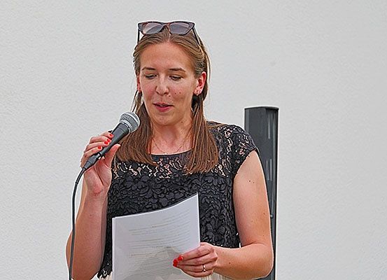 Eine Frau spricht in ein Mikrofon und hält ein Stück Papier in der anderen Hand. Sie trägt ein schwarzes Spitzenoberteil und eine Sonnenbrille auf dem Kopf.