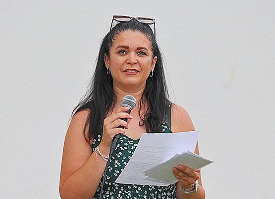 Eine Frau hält ein Papier und ein Mikrofon mit einem Lächeln, möglicherweise beim Lesen oder Sprechen. Sie trägt ein ärmelloses Blumenkleid.