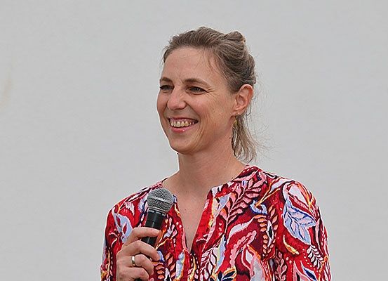Eine Frau mit kurzen Haaren hält ein Mikrofon in der Hand und lächelt, wahrscheinlich bei einer Veranstaltung im Freien. Sie trägt ein rotes Blumenblusen und einen Ring.