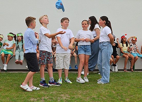 Gruppe von Kindern in Kostümen mit Stirnbändern steht auf einem Rasenfeld, wahrscheinlich bei einer Aufführung. Ein blauer Hut ist in der Luft. Einige sitzen auf einer Bank hinter ihnen.