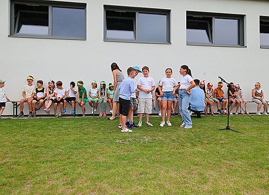 Eine Gruppe von Kindern und Erwachsenen ist vor einem Gebäude versammelt. Sie sitzen auf Bänken, und einige stehen davor. Die Kinder tragen Hüte und Turnschuhe. Ein Mikrofon steht auf dem Rasen.