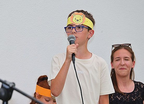 Ein junger Junge mit einem Löwen-Kopfband hält ein Mikrofon und spricht hinein. Eine Frau mit Sonnenbrille auf dem Kopf schaut hinter ihm nach unten. Ein weiteres Kind mit einem orangefarbenen Kopfband ist teilweise im Hintergrund zu sehen.