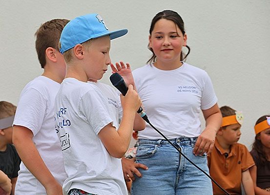 Ein junger Junge mit Mikrofon steht vor einer Gruppe von Kindern, die weiße T-Shirts, blaue Jeans und Stirnbänder mit Eulendesign tragen. Hinter ihm steht ein Mädchen, das ebenfalls ein weißes T-Shirt trägt. Sie stehen vor einer weißen Wand.