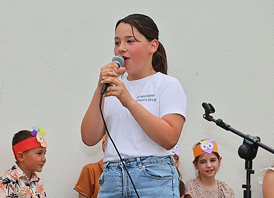 Ein Mädchen singt in ein Mikrofon bei einer Veranstaltung im Freien, trägt ein weißes T-Shirt, mit Kindern im Hintergrund, die Stirnbänder tragen.