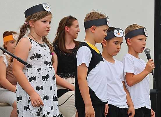 Eine Gruppe von Kindern mit Pinguin-Kopfbändern, eines hält ein Mikrofon, steht zusammen mit einer Frau im Hintergrund.