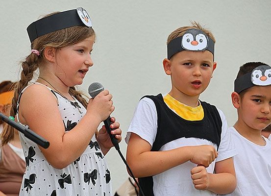 Drei Kinder führen auf, ein Mädchen und zwei Jungen, mit Pinguinkopfbändern. Das Mädchen hält ein Mikrofon, während die Jungen in die Kamera schauen.