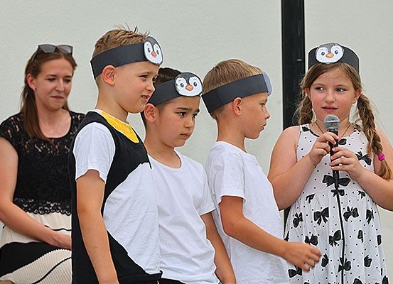 Fünf Kinder mit Pinguinkopfbändern stehen in einer Reihe, eine Mädchen hält ein Mikrofon. Dahinter beobachtet eine Frau mit Sonnenbrille auf dem Kopf.
