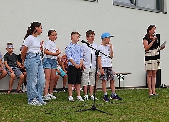 Eine Gruppe von Kindern in Sommerkleidung steht vor einem Mikrofon, während eine Frau aus einem Papier liest. Hinter ihnen sitzen Kinder auf einer Bank, und ein Gebäude ist im Hintergrund.