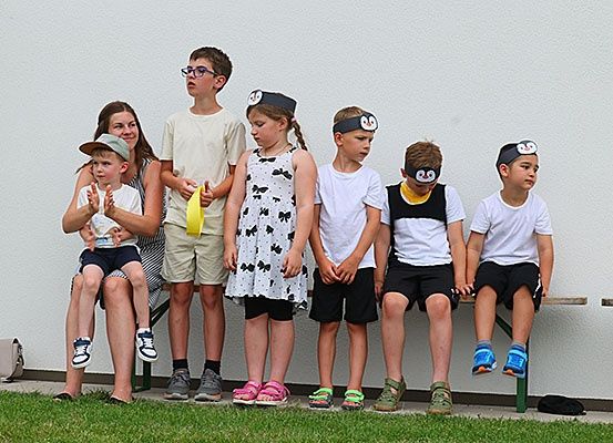 Eine Gruppe von Kindern mit Stirnbändern sitzt auf einer Bank. Ein Kind hält ein gelbes Band. Eine Frau hält ein Kind auf dem Schoß. Sie alle schauen nach vorne.