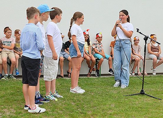 Eine Gruppe von Kindern steht und sitzt auf einer Bank. Ein Mädchen hält ein Mikrofon und scheint zu singen. Hinter ihr beobachten einige Kinder sie. Der Boden ist mit Gras bedeckt.