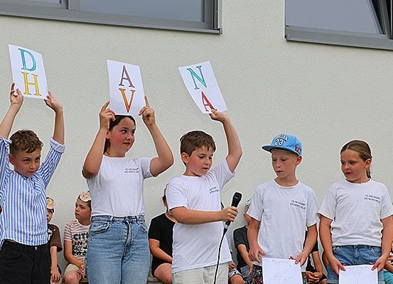 Kinder halten Karten mit Buchstaben hoch, während ein Junge ein Mikrofon in der Hand hat. Sie stehen vor einem Gebäude.