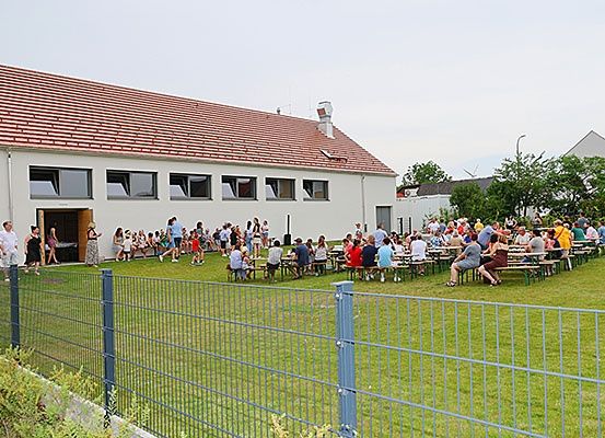 Eine Gruppe von Menschen versammelt sich vor einem weißen Gebäude mit rotem Dach und sitzt an Tischen und unterhält sich. Das Gebäude hat mehrere Fenster und eine Tür. Ein Zaun umgibt den Rasenbereich. Bäume und andere Gebäude sind in der Ferne sichtbar.