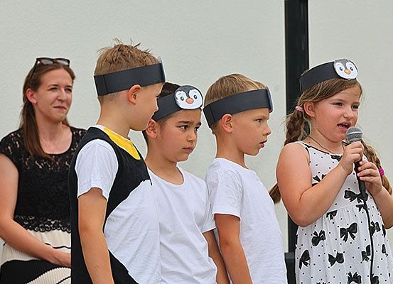 Vier Kinder mit Pinguin-Kopfbändern stehen zusammen, eines hält ein Mikrofon. Eine Frau mit Sonnenbrille auf dem Kopf beobachtet sie von hinten.