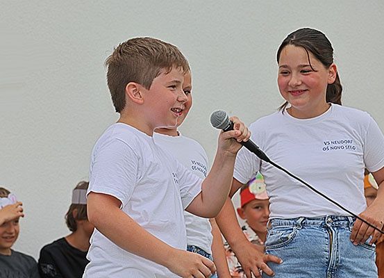 Eine Gruppe von Kindern in weißen T-Shirts tritt auf der Bühne mit einem Mikrofon auf. Ein junger Junge hält das Mikrofon, und ein Mädchen neben ihm lächelt. Hinter ihnen beobachten andere Kinder. Die Wand im Hintergrund ist weiß.