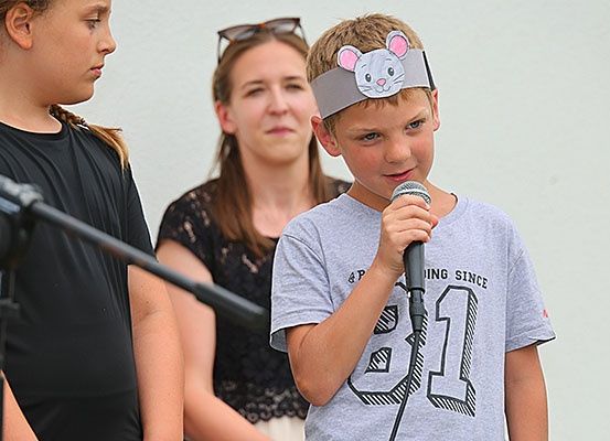 Ein junger Junge mit einer Maus-Kopfband spricht in ein Mikrofon, während zwei Frauen hinter ihm stehen, eine mit einer Brille auf dem Kopf.