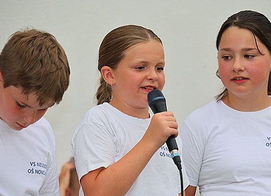 Drei Kinder, zwei Mädchen und ein Junge, stehen eng beieinander, wobei das Mädchen in der Mitte ein Mikrofon hält. Sie tragen passende weiße T-Shirts.